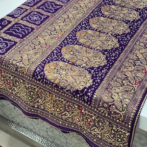 Pu&ograve; includere: Tessuto viola con intricati motivi dorati e bianchi. Il design include forme geometriche, motivi paisley e accenti a pois. Il tessuto ha un bordo dettagliato con elementi floreali e simili ad uccelli.