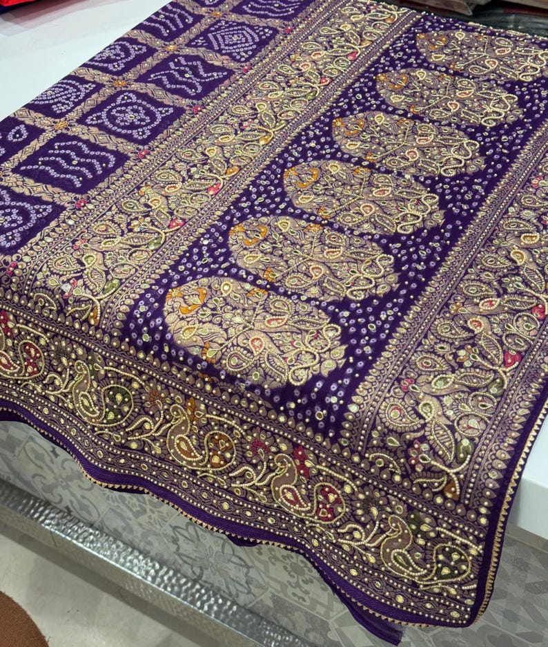 Pu&ograve; includere: Tessuto viola con intricati motivi dorati e bianchi. Il design include forme geometriche, motivi paisley e accenti a pois. Il tessuto ha un bordo dettagliato con elementi floreali e simili ad uccelli.