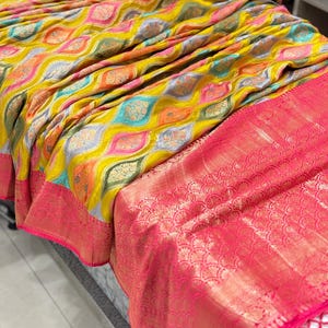 May include: A yellow, pink, and blue patterned silk saree with a pink border and a gold trim. The saree has a decorative fringe.