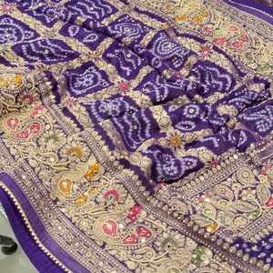 Pu&ograve; includere: Primo piano di un tessuto viola con un motivo dettagliato. Il tessuto presenta un motivo quadrato ripetuto e intricati motivi floreali e paisley in oro, rosa e verde. I bordi sono rifiniti con un bordo decorativo.