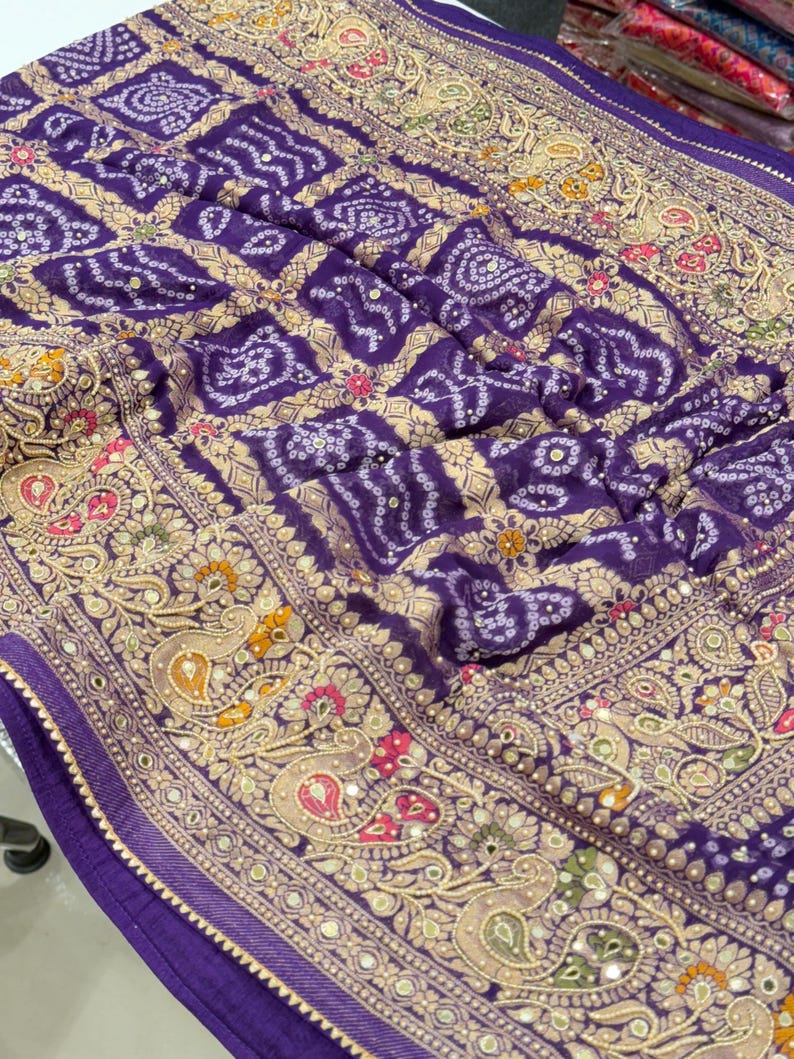 Pu&ograve; includere: Primo piano di un tessuto viola con un motivo dettagliato. Il tessuto presenta un motivo quadrato ripetuto e intricati motivi floreali e paisley in oro, rosa e verde. I bordi sono rifiniti con un bordo decorativo.