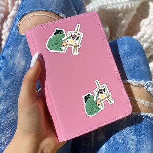 May include: Two frog stickers on a pink notebook. Each frog is wearing a beret and holding a paintbrush. The frogs are painting flowers on a canvas.