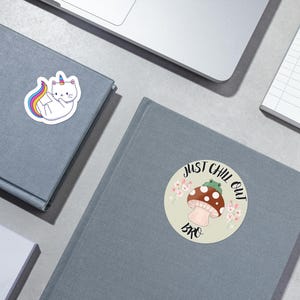 May include: Two stickers on a grey surface. One sticker features a white cat with a rainbow tail and a unicorn horn. The other sticker features a mushroom with a green frog on top and the text "Just Chill Out Bro".