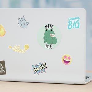 May include: A laptop computer with various colourful stickers, including a frog with the text "Bite Me", a yellow star with pink eyes, a light blue sticker with the text "Dream Big", a yellow lightbulb with the text "Idea", a brown sticker with the text "Coffee is always a good idea", a yellow sticker with the text "Boss!" and a grey cat with a white face.