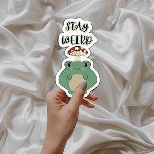 May include: A green frog sticker with a red and white mushroom on its head. The sticker says "Stay Weird" in black text.