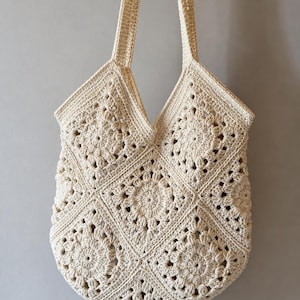 May include: Cream-colored crochet tote bag with two long straps. The bag features a floral pattern created with a series of squares. The bag is made of a soft, textured yarn and is suitable for carrying various items.