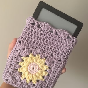 以下が含まれることがあります： ラベンダー色の手編みKindleケース。黄色とピンクのヒマワリのデザイン。手作りのケースはスカラップエッジとボタン留めが特徴です。Kindleは黒色です。