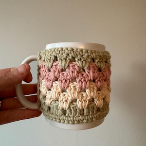May include: A white ceramic mug with a crocheted cozy in shades of green, cream, and pink. The cozy is made with a repeating pattern of small squares.