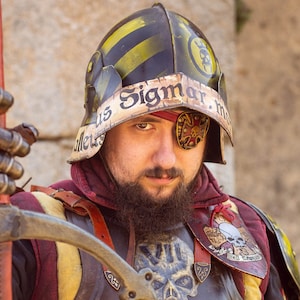 May include: A person wearing a yellow and black helmet with the text "Sigmar" on it, a red and gold tunic, and a brown leather breastplate with a skull and crossbones design. The person is holding a long, wooden staff with a metal tip.