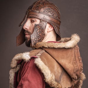 K&ouml;nnte beinhalten: Braunes Leder-Wikinger-Kriegerkost&uuml;m mit Helm, fellgef&uuml;ttertem Umhang und detailliertem G&uuml;rtel. Das Kost&uuml;m beinhaltet ein Schwert und eine Scheide, die ein historisches und Fantasy-Thema zeigen. Das Bild zeigt das Logo "El Marqu&eacute;s LARP Shop".