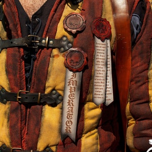 Könnte beinhalten: Nahaufnahme einer gelbschwarzen gestreiften Weste mit Lederriemen und Schnallen. An der Weste sind drei Wachssiegel befestigt, jedes mit einem anderen Design. Die Siegel sind an Bändern befestigt, auf denen Text geschrieben steht. Der Text auf den Bändern lautet "Imperator" und "Das Kreuz mit einer Pilgerreise ins Heilige Land".