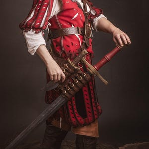 Op de afbeelding: Een persoon in een rood en wit middeleeuws kostuum, met een zwaard. Het kostuum omvat een rode wambuis met witte mouwen, een bruine riem en zwarte laarzen. Het zwaard heeft een rode handgreep en een zilveren lemmet.