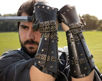 May include: A pair of black leather arm guards with brass accents and a laced design. The guards are worn on both arms and have a medieval or fantasy aesthetic.