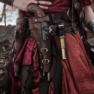 Puede incluir: Una mujer con un vestido rojo con un cintur&oacute;n de cuero marr&oacute;n y dos pistolas en fundas en sus caderas. Las pistolas est&aacute;n en fundas de cuero marr&oacute;n con detalles met&aacute;licos. La mujer tambi&eacute;n lleva una pulsera de plata en su mu&ntilde;eca derecha.