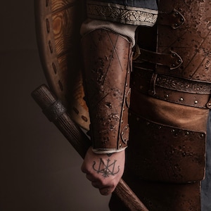 May include: A person wearing brown leather armor with a shield and an axe. The axe has a wooden handle and a metal head. The person has a tattoo on their hand.