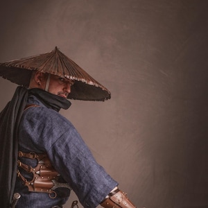 May include: A person wearing a blue shirt and brown leather armor with a wide-brimmed straw hat, holding a sword and crouching on a brown background.