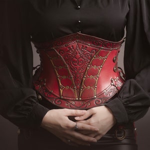 K&ouml;nnte beinhalten: Ein rotes Lederkorsett mit aufwendigen goldenen und schwarzen Details. Das Korsett hat eine spitze Vorderseite und wird mit Metallverschl&uuml;ssen geschlossen.