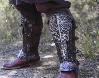 May include: Close-up of a pair of brown leather boots with silver metal armor plates on the shins. The armor plates have intricate designs and are secured with buckles.