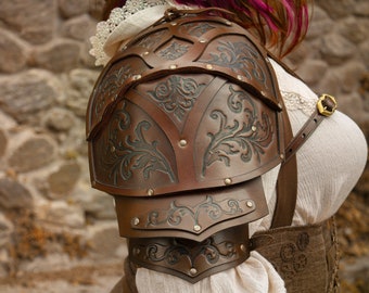Puede incluir: Hombreras de cuero marrón con intrincados diseños florales y remaches de latón. La armadura forma parte de un disfraz, con placas superpuestas y una correa con hebilla dorada. El diseño sugiere un tema medieval o de fantasía.
