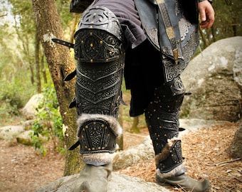 Peut inclure: Armure de jambe de style médiéval noir et argenté avec des motifs en relief complexes et une garniture en fourrure. L'armure comporte de multiples sangles et rivets. L'image est prise dans un environnement extérieur naturel.