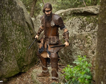 May include: A man dressed in brown leather armor with a fur-trimmed cloak and a braided beard, holding two swords in a forest setting.