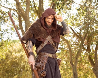May include: A man wearing a brown hooded cloak with fur trim, a leather belt with pouches, and a staff. He is smiling and looking at the camera.