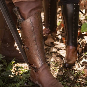 May include: Close-up of a brown leather boot with metal accents and buckles. The boot is worn and has a distressed look. The boot is on a dirt path with green plants and brown leaves.