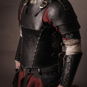 Peut inclure: Une armure en cuir noir avec des accents en laiton décoratifs. L'armure comprend une pièce de poitrine, des épaulettes et des protège-bras. L'armure est portée par une personne avec une barbe et une chemise rouge.