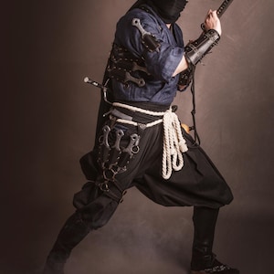 May include: A person dressed as a ninja wearing a black mask, a brown conical hat, and a blue shirt. They are holding a sword and have a variety of throwing knives strapped to their legs.