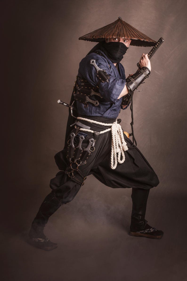 May include: A person dressed as a ninja wearing a black mask, a brown conical hat, and a blue shirt. They are holding a sword and have a variety of throwing knives strapped to their legs.