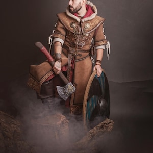 Peut inclure: Une personne porte un costume de Viking en cuir marron, tenant une hache et un bouclier. Le costume comprend une capuche doubl&eacute;e de fourrure, des d&eacute;tails complexes et un sous-v&ecirc;tement rouge. La hache a un manche rouge et une t&ecirc;te argent&eacute;e. Le bouclier est bleu et noir.