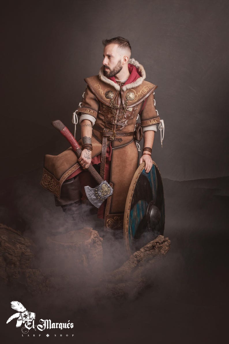 Peut inclure: Une personne porte un costume de Viking en cuir marron, tenant une hache et un bouclier. Le costume comprend une capuche doubl&eacute;e de fourrure, des d&eacute;tails complexes et un sous-v&ecirc;tement rouge. La hache a un manche rouge et une t&ecirc;te argent&eacute;e. Le bouclier est bleu et noir.