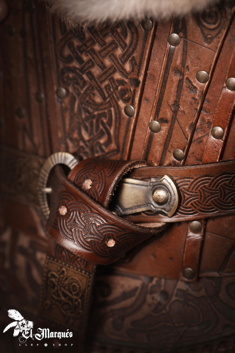 May include: Close-up of a brown leather belt with intricate Celtic knot designs. The belt has a metal buckle with a decorative design and is fastened with a metal rivet.