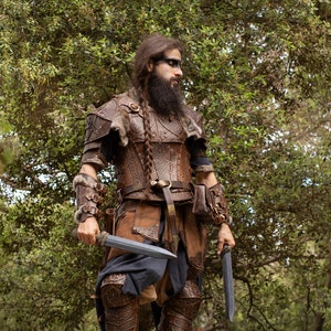 May include: A man dressed in a brown leather costume with a long braid and beard, holding two knives in his hands. He is standing in a forest setting.