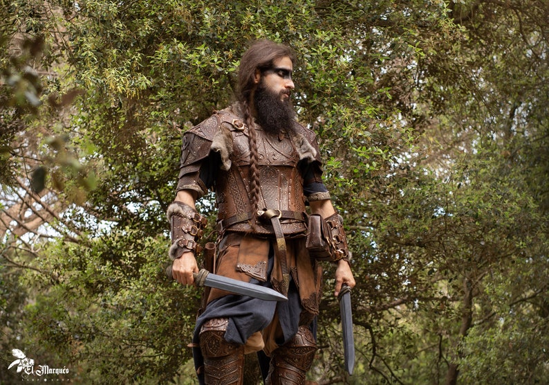 May include: A man dressed in a brown leather costume with a long braid and beard, holding two knives in his hands. He is standing in a forest setting.