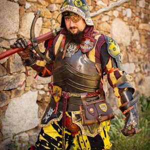 K&ouml;nnte beinhalten: Eine Person in mittelalterlicher R&uuml;stung mit gelber, schwarzer und roter Farbgebung. Die R&uuml;stung umfasst einen Helm mit Totenkopf- und Knochenkreuz-Design, einen Brustpanzer und Handschuhe. Die Person h&auml;lt ein Schwert mit einer gebogenen Klinge.