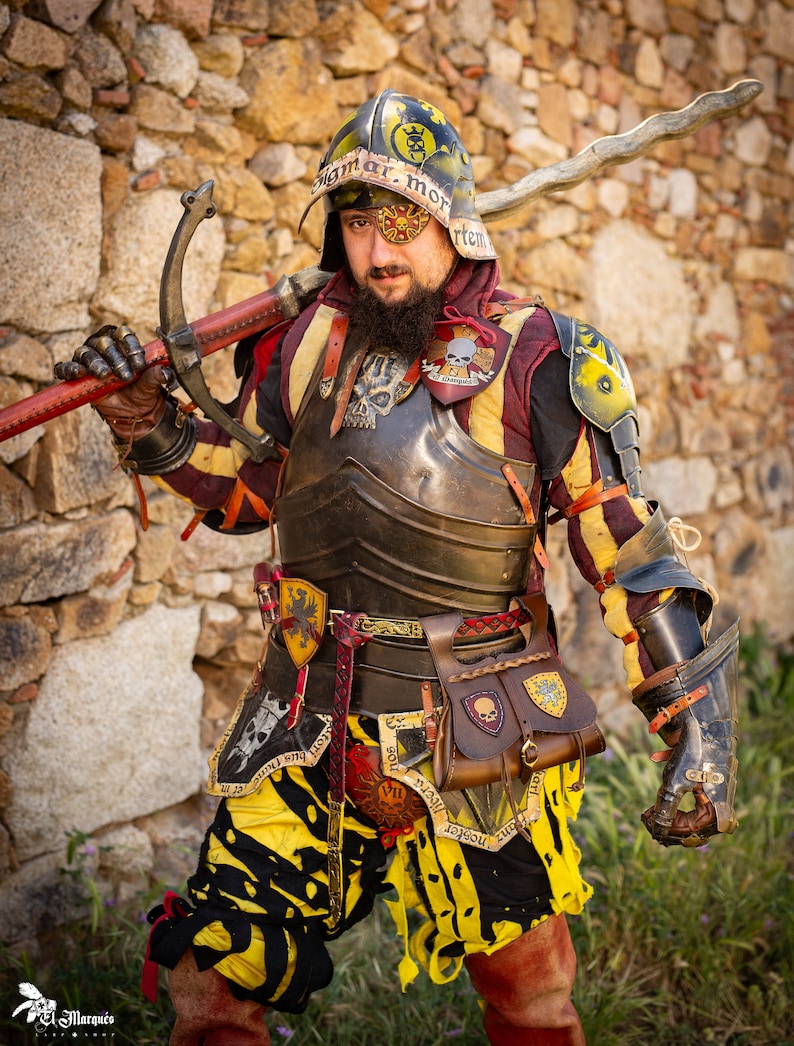 K&ouml;nnte beinhalten: Eine Person in mittelalterlicher R&uuml;stung mit gelber, schwarzer und roter Farbgebung. Die R&uuml;stung umfasst einen Helm mit Totenkopf- und Knochenkreuz-Design, einen Brustpanzer und Handschuhe. Die Person h&auml;lt ein Schwert mit einer gebogenen Klinge.