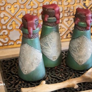 May include: Three small green glass bottles with a white label and a red wax seal on the top. The bottles are sitting on a black metal tray with a wooden spoon.