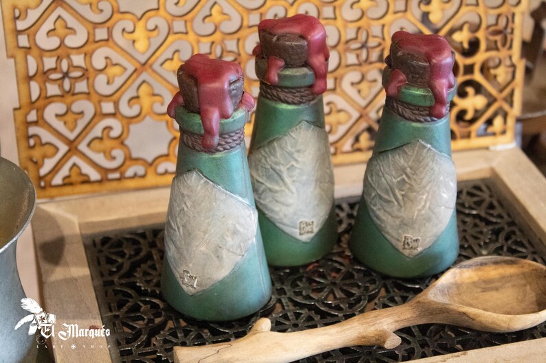 May include: Three small green glass bottles with a white label and a red wax seal on the top. The bottles are sitting on a black metal tray with a wooden spoon.