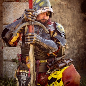 K&ouml;nnte beinhalten: Eine Person in mittelalterlicher R&uuml;stung und einem Helm mit gelbem und schwarzem Streifenmuster. Die Person h&auml;lt ein Schwert und hat ein Totenkopf- und Knochenkreuz-Emblem auf der Brust. Der Text "us Sigm" ist auf dem Helm sichtbar.