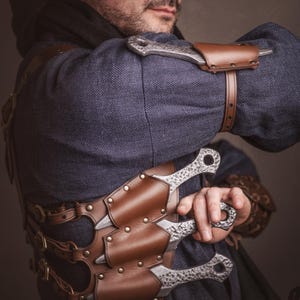 May include: A person wearing a brown leather chest rig with multiple silver throwing knives. The rig has straps and buckles. The person is wearing a blue shirt and a brown hat.