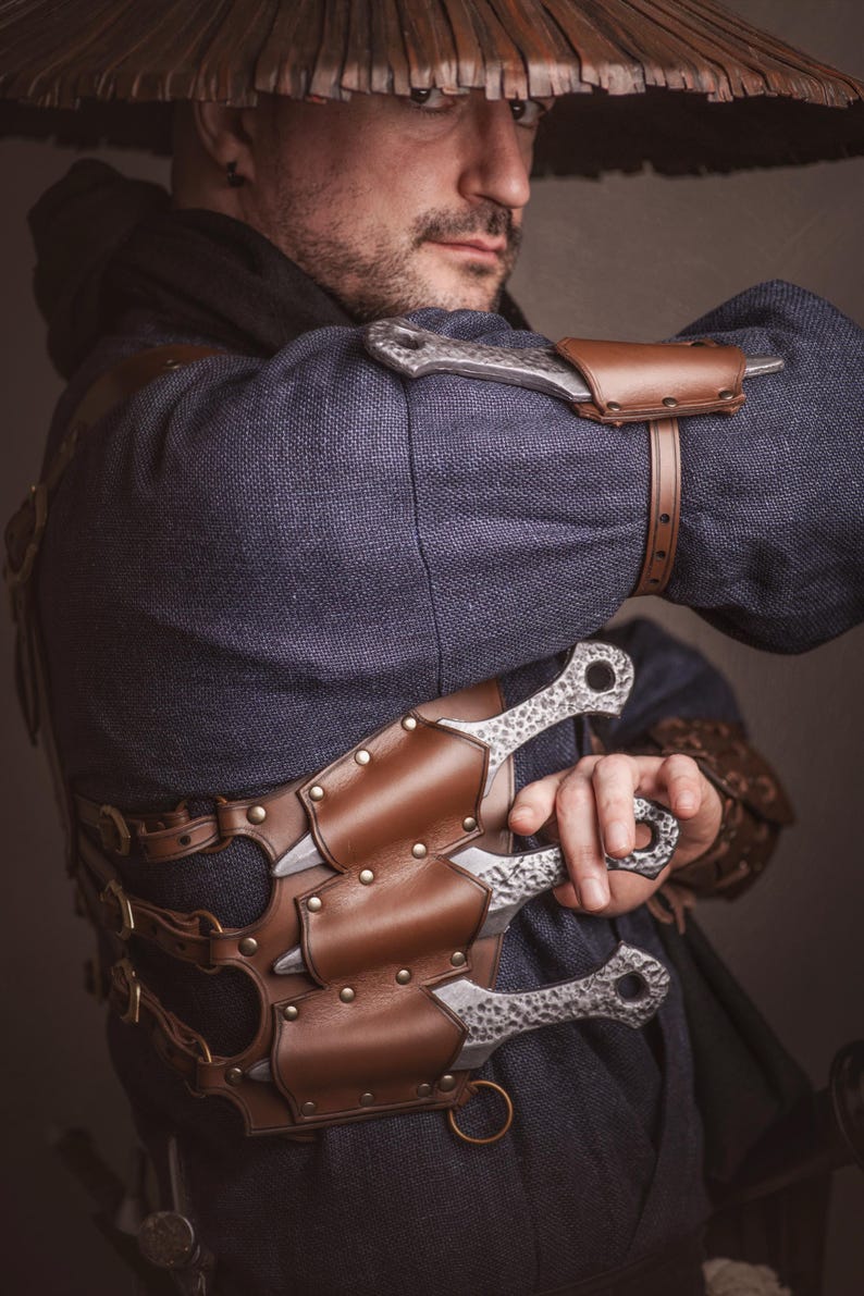 May include: A person wearing a brown leather chest rig with multiple silver throwing knives. The rig has straps and buckles. The person is wearing a blue shirt and a brown hat.