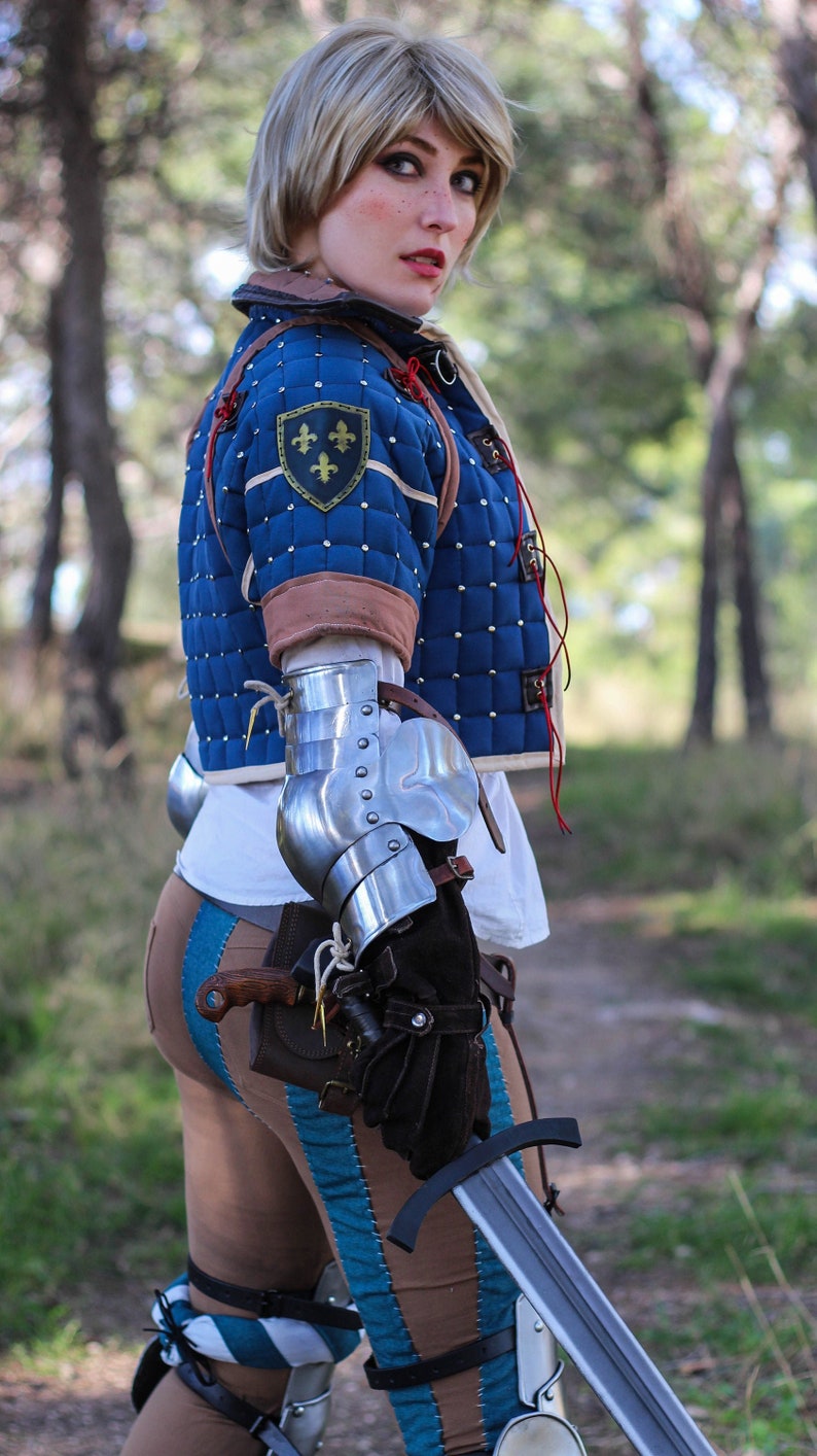 K&ouml;nnte beinhalten: Eine Frau in einer blauen und braunen gesteppten Jacke mit einem goldenen Fleur-de-lis-Emblem, die ein Schwert h&auml;lt und eine Metallr&uuml;stung am Arm tr&auml;gt.