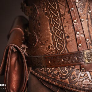 May include: A brown leather corset with intricate knotwork and metal accents. The corset has a belt with a decorative buckle and a small leather pouch attached to the side.