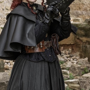 May include: A woman in a black costume with a long black cape and a black hat with a feather. She is holding a black plague doctor mask with a long beak. The mask has a leather strap and is decorated with metal studs. The woman is standing in front of a stone wall.
