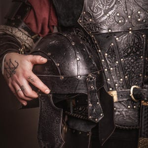 K&ouml;nnte beinhalten: Schwarzer Lederhelm mit genieteten Akzenten, gehalten vor einer passenden schwarzen Lederbrustplatte. Die R&uuml;stung weist aufwendige gepr&auml;gte Designs und goldfarbene Schnallen auf. Das Bild zeigt mittelalterliche Kleidung.