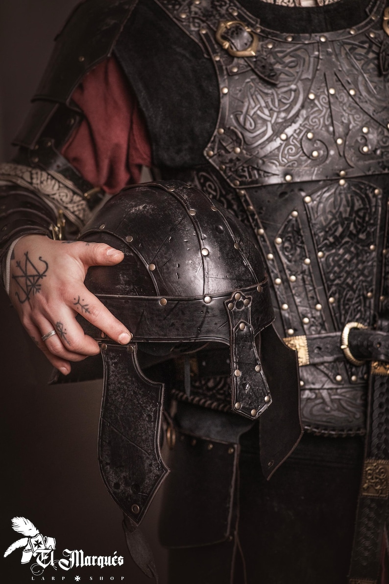 K&ouml;nnte beinhalten: Schwarzer Lederhelm mit genieteten Akzenten, gehalten vor einer passenden schwarzen Lederbrustplatte. Die R&uuml;stung weist aufwendige gepr&auml;gte Designs und goldfarbene Schnallen auf. Das Bild zeigt mittelalterliche Kleidung.