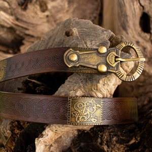 May include: A brown leather belt with a brass buckle and decorative metal accents. The belt has a Celtic knot design and a square metal plate with a stylized animal design.