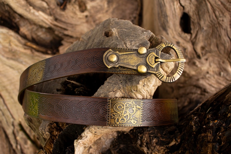 May include: A brown leather belt with a brass buckle and decorative metal accents. The belt has a Celtic knot design and a square metal plate with a stylized animal design.
