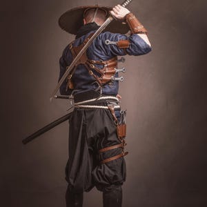 May include: A person wearing a blue shirt and black pants with a wide-brimmed hat, holding a sword over their shoulder. They are wearing a leather harness with multiple knives attached.